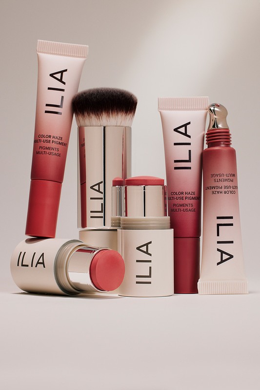 Stacked Ilia makeup products on a white surface: cream blush, bronzer stick, makeup brush, and multi-use pigment tubes, studio shot.