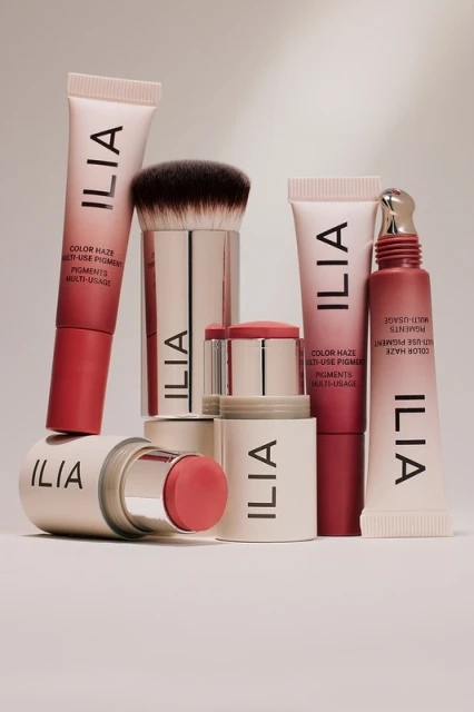 Stacked Ilia makeup products on a white surface: cream blush, bronzer stick, makeup brush, and multi-use pigment tubes, studio shot.