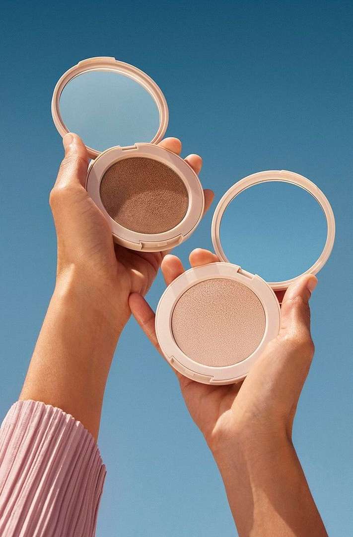 Woman presents two round makeup compacts against a clear blue sky, highlighting beauty product presentation and natural outdoor lighting.