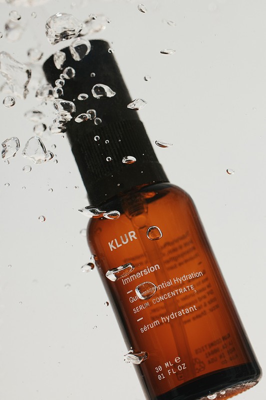 A glass amber bottle of KLUR serum concentrate suspended underwater with water bubbles surrounding it, set against a white background.