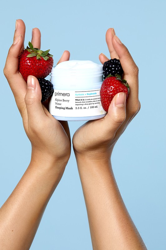 Two hands hold a jar of Primera Alpine Berry Sleeping Mask surrounded by strawberries and blackberries on a light blue background.