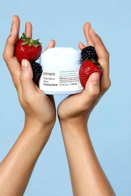 Two hands hold a jar of Primera Alpine Berry Sleeping Mask surrounded by strawberries and blackberries on a light blue background.