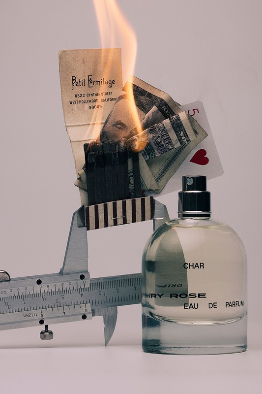 Perfume bottle next to a caliper tool, burning paper, dollar bill, and playing card on a white background in a studio setup.