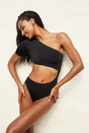 Smiling model wearing a black one-shoulder cutout swimsuit against a light neutral textured background.