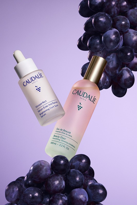 Two Caudalie skincare product bottles are surrounded by a pile and crown of dark purple grapes against a soft purple backdrop.