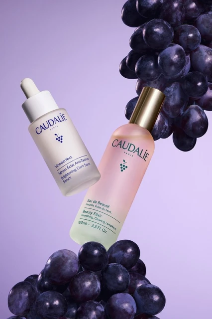 Two Caudalie skincare product bottles are surrounded by a pile and crown of dark purple grapes against a soft purple backdrop.