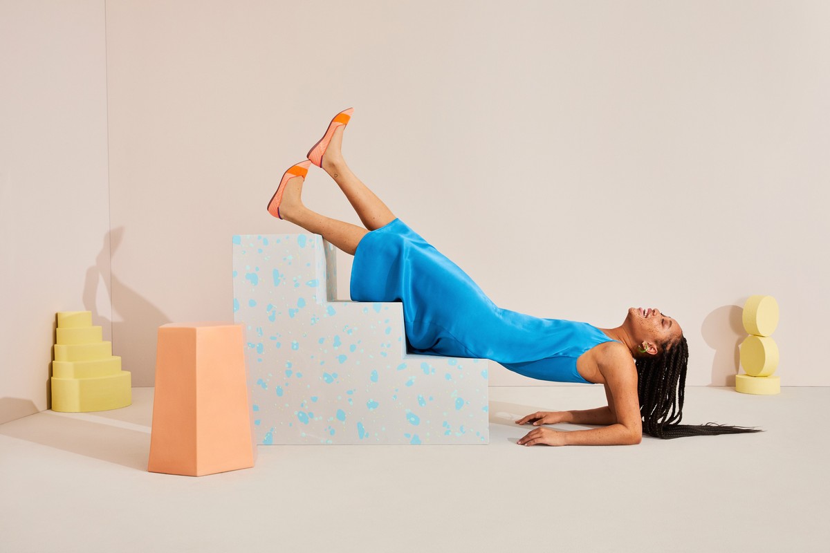 Woman in blue dress and orange shoes poses on geometric steps in a studio with pastel yellow and peach props, lying on her back, smiling.