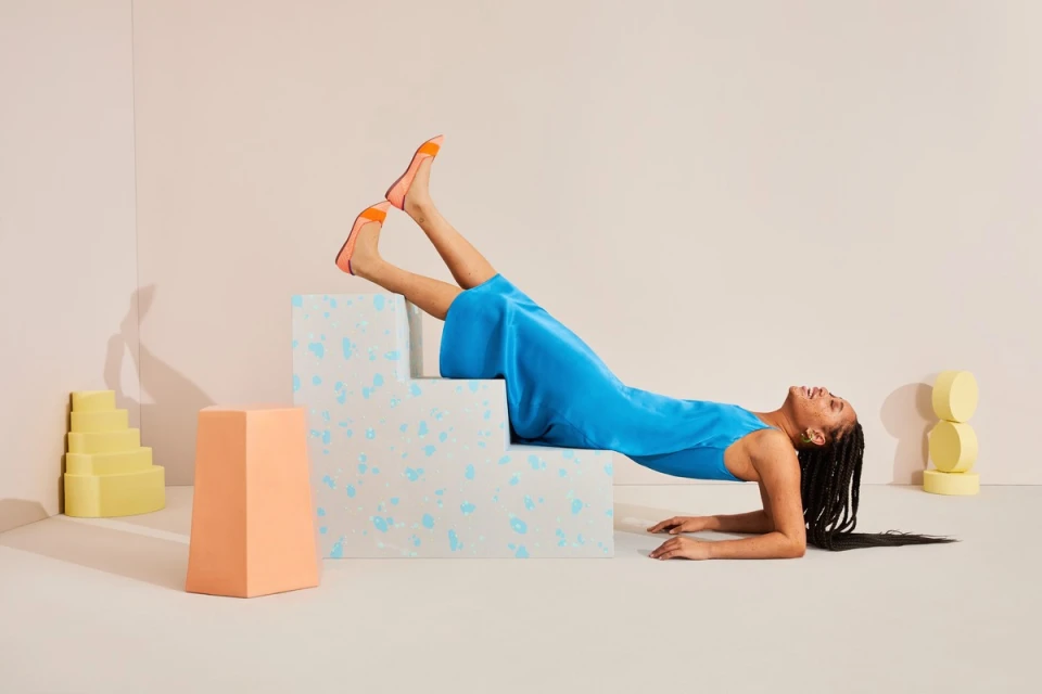 Woman in blue dress and orange shoes poses on geometric steps in a studio with pastel yellow and peach props, lying on her back, smiling.