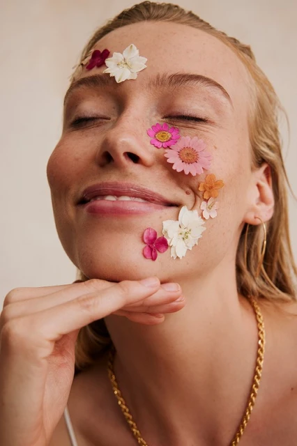 Close-up portrait of a smiling woman with closed eyes and colorful pressed flowers adhered to her face and gold chain necklace.