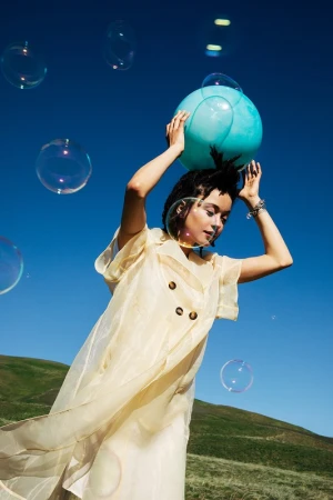 Woman in a sheer yellow coat holding a turquoise ball overhead outdoors with floating bubbles against a clear blue sky