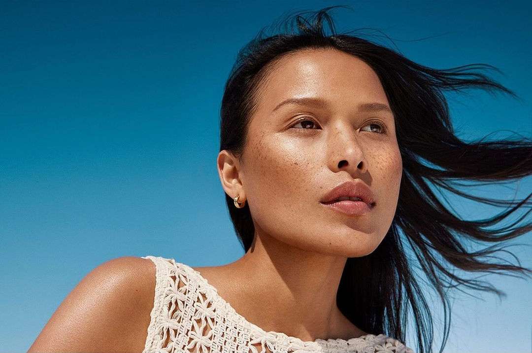 Close-up of an adult woman with an upward gaze, showcasing her skin texture and freckles against a clear blue sky backdrop.