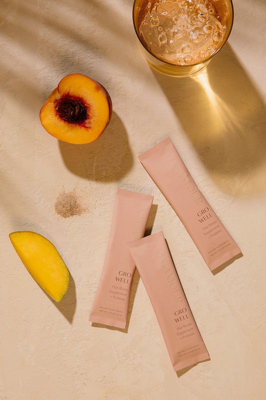 Overhead shot of peach halves, mango slices, and Grow Well supplement packets arranged with a glass of drink on a beige surface.