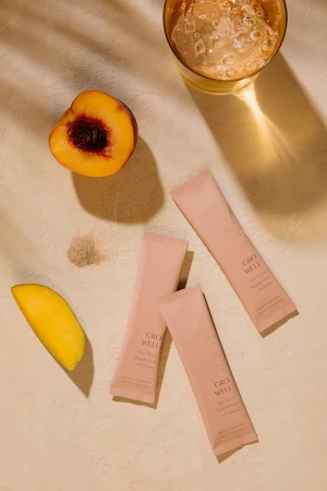 Overhead shot of peach halves, mango slices, and Grow Well supplement packets arranged with a glass of drink on a beige surface.