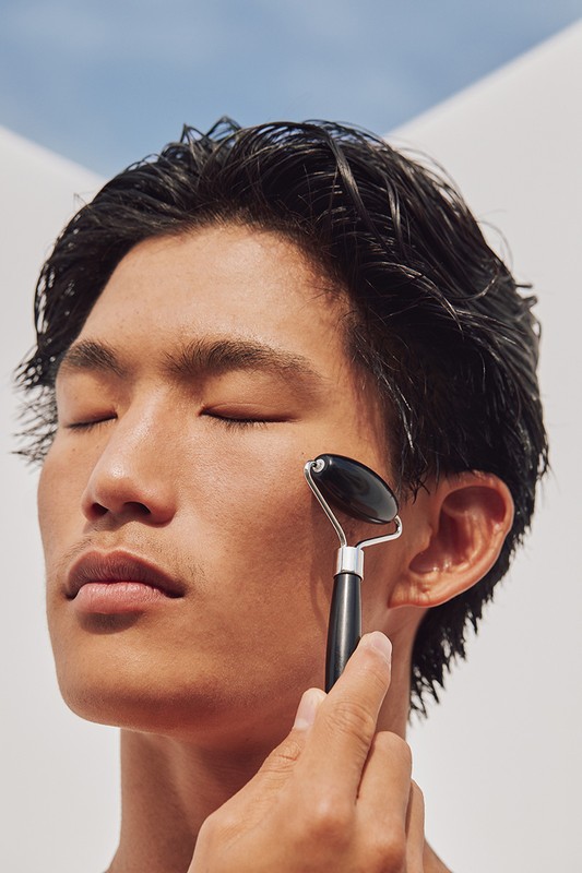 Close-up of an Asian man with dark hair using a black facial roller on his face against a white and blue background in soft light.