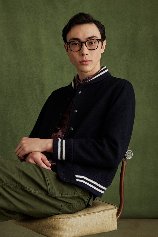 Portrait of a young adult male sitting on a chair wearing a letterman jacket, olive-green pants and glasses against a green backdrop.