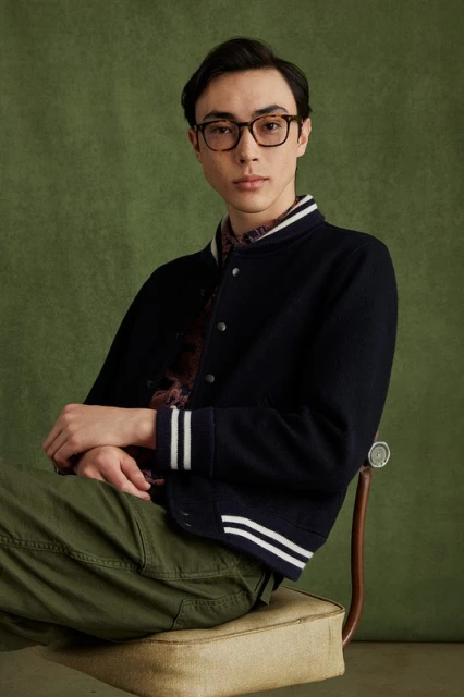 Portrait of a young adult male sitting on a chair wearing a letterman jacket, olive-green pants and glasses against a green backdrop.