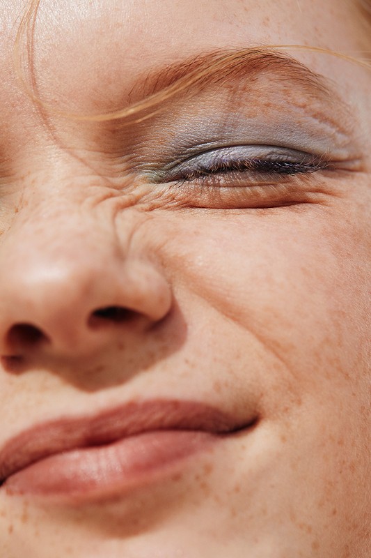 Close-up portrait of a woman with freckles and blue eyeshadow, squinting her eyes slightly in bright sunlight, showcasing beauty details.