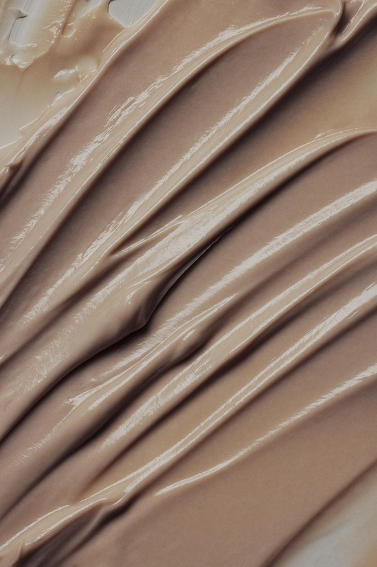 Close-up of a beige-colored cream or cosmetic product, showing smooth, even strokes and a reflective, glossy texture.