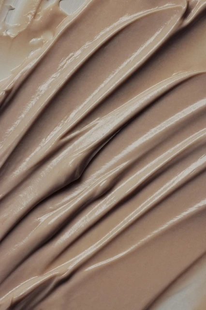 Close-up of a beige-colored cream or cosmetic product, showing smooth, even strokes and a reflective, glossy texture.
