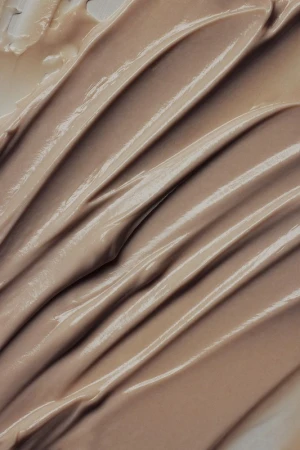Close-up of a beige-colored cream or cosmetic product, showing smooth, even strokes and a reflective, glossy texture.