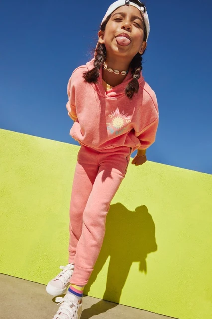 A young girl sticks her tongue out at the camera. She is wearing a pink sweat suit and a white baseball cap while leaning on a chartreuse wall.
