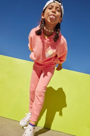 A young girl sticks her tongue out at the camera. She is wearing a pink sweat suit and a white baseball cap while leaning on a chartreuse wall.