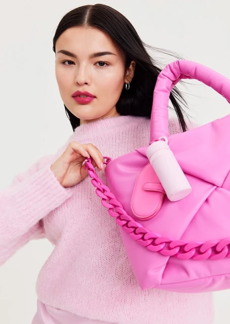A young woman poses with a pink, quilted, puffy handbag with chain detail, wearing a matching pink sweater and skirt against a white background.