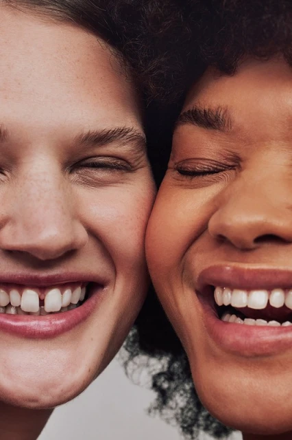 Close-up portrait of two smiling faces: a Caucasian female with a gap-toothed smile and an African-American woman laughing joyfully.