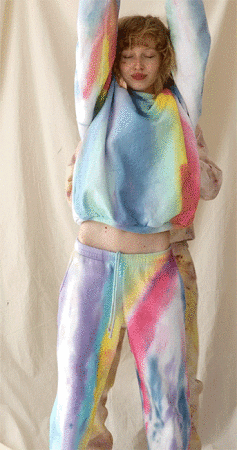 A woman with curly hair poses with arms raised wearing a colorful pastel-tone tie-dye sweat suit, set against an off-white background.