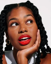 Close-up portrait of a woman with red lipstick and braided black hair resting her hand on her chin against a white background.