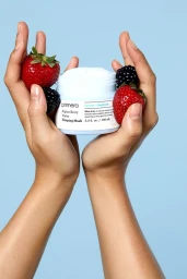 Two hands holding a skincare jar surrounded by fresh strawberries and blackberries against blue background.
