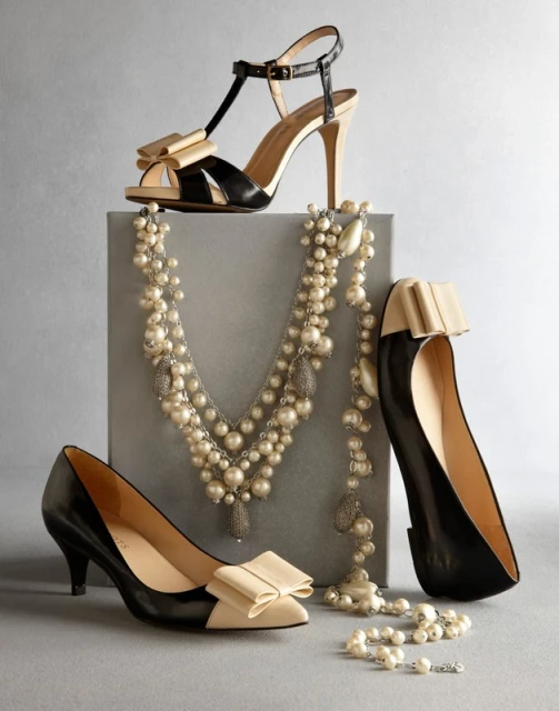Black leather high heel shoes and a pearl necklace are arranged on a grey block and background in a studio product shot.