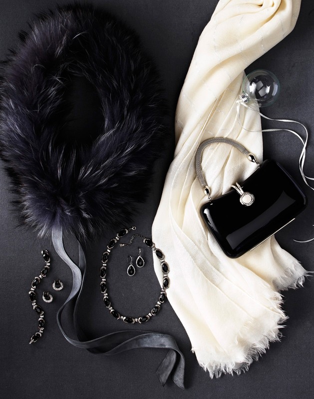 Arrangement of women's fashion accessories: fur collar, black purse, pearl necklace, jewelry set and white scarf on dark gray background.