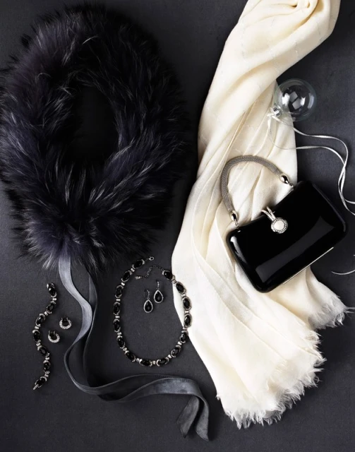 Arrangement of women's fashion accessories: fur collar, black purse, pearl necklace, jewelry set and white scarf on dark gray background.