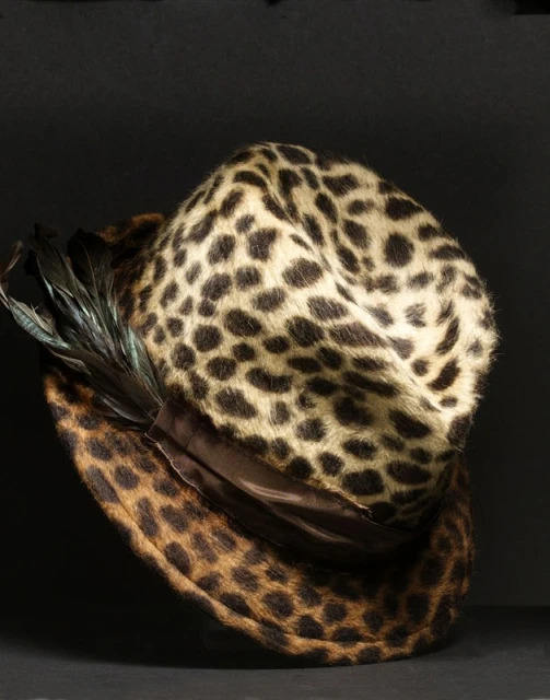 Leopard print fedora hat with a brown band and black feathers displayed against a dark gray background in a studio product shot.