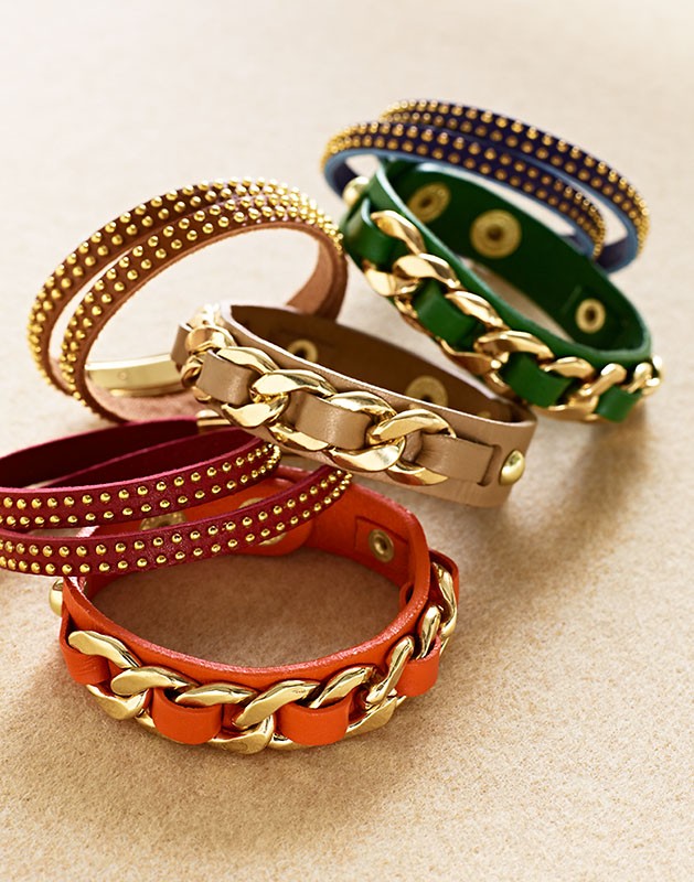 Close-up of a colorful collection of leather and chain bracelets with gold hardware, arranged on a neutral surface, studio lighting.