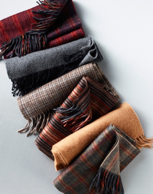 An arrangement of folded wool scarves in various plaid, striped, and solid patterns featuring fringe details against a light grey background.