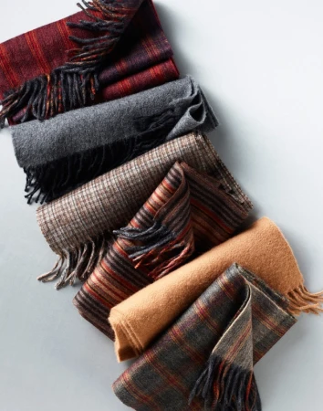 An arrangement of folded wool scarves in various plaid, striped, and solid patterns featuring fringe details against a light grey background.