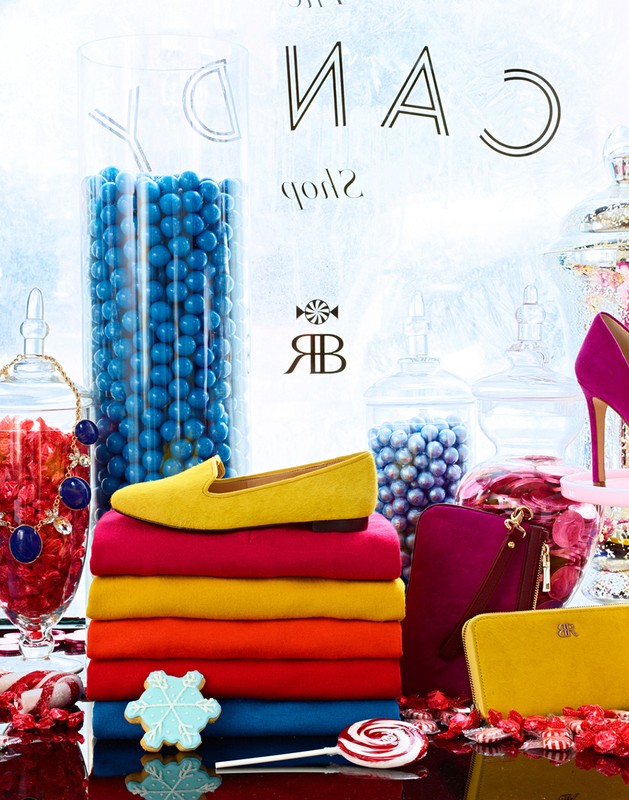 Festive flat lay features a yellow loafer, stacked sweaters, handbags, candy-filled jars, and holiday treats in a bright, colourful composition.