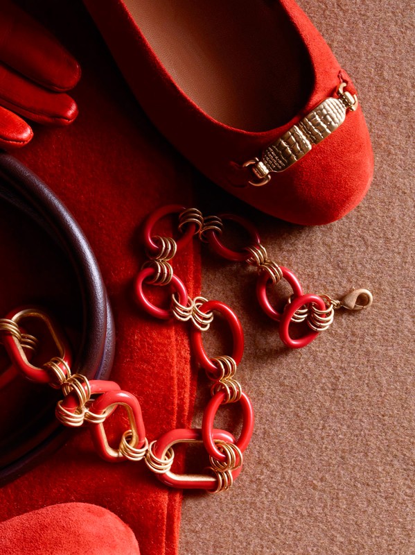 Still life featuring a red ballet flat with a gold embellishment, a red linked bracelet, a red glove and purse on a beige surface.