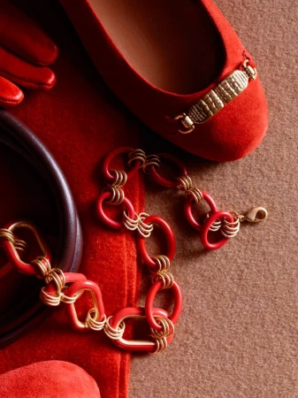 Still life featuring a red ballet flat with a gold embellishment, a red linked bracelet, a red glove and purse on a beige surface.