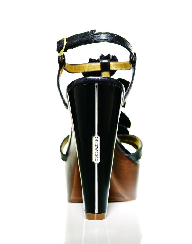 Elevated view of a black leather high-heeled Coach sandal with a floral accent, displayed on a white background. The platform sole is made of wood.