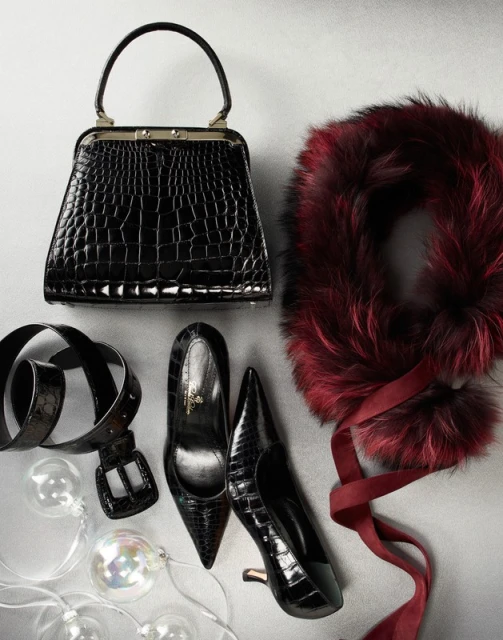 Fashion still life featuring a black crocodile leather handbag, black pumps, a belt, a faux fur scarf with ribbon, and decorative lights.