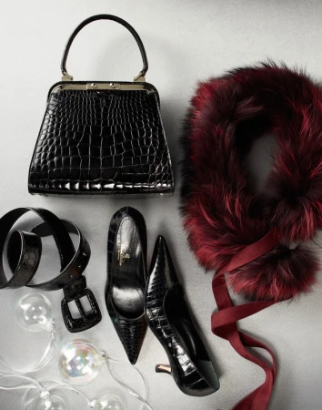 Fashion still life featuring a black crocodile leather handbag, black pumps, a belt, a faux fur scarf with ribbon, and decorative lights.