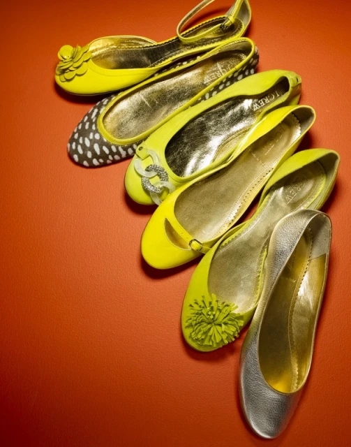 Arrangement of several pairs of women's ballet flat shoes in shades of yellow and metallic silver on a textured coral background.