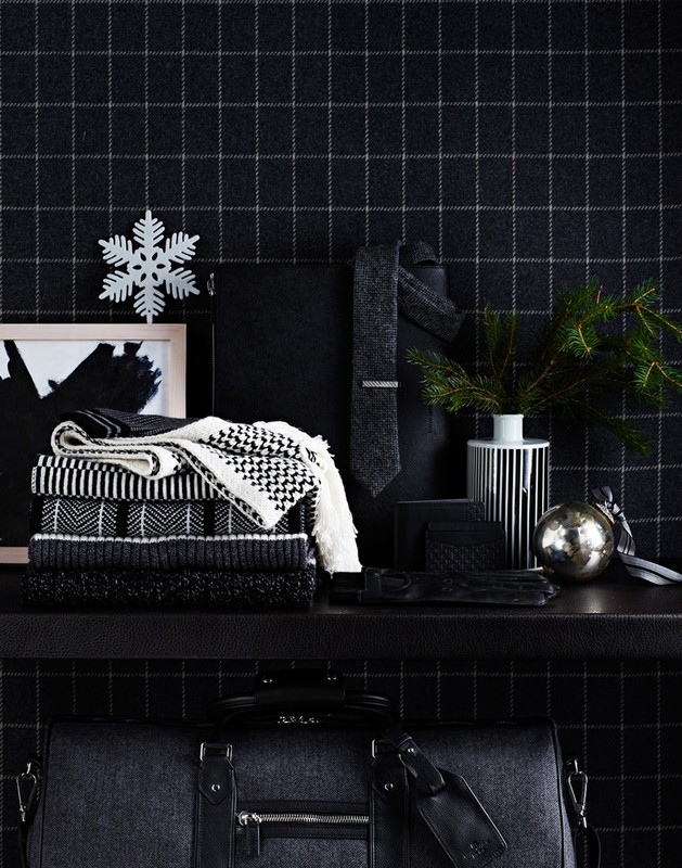 Stacked sweaters, a tie, gloves, an ornament, and a duffel bag on a shelf against a black and gray plaid wallpaper backdrop.
