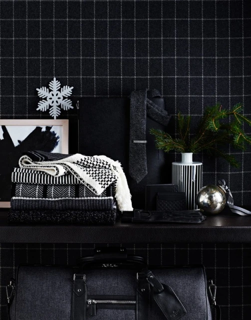 Stacked sweaters, a tie, gloves, an ornament, and a duffel bag on a shelf against a black and gray plaid wallpaper backdrop.