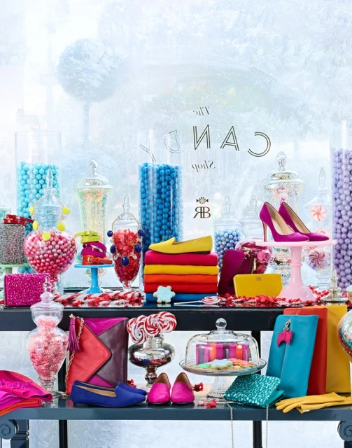Display of colorful shoes, handbags, and candy in jars, arranged on a black table against a pale blue, abstract background, product photography.