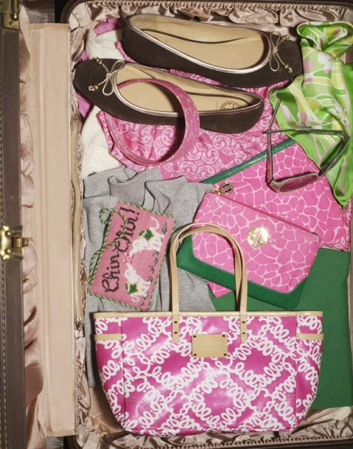 Open suitcase filled with pink patterned tote bag, brown ballet flats, various clothing items, eyeglasses, and accessories for travel.