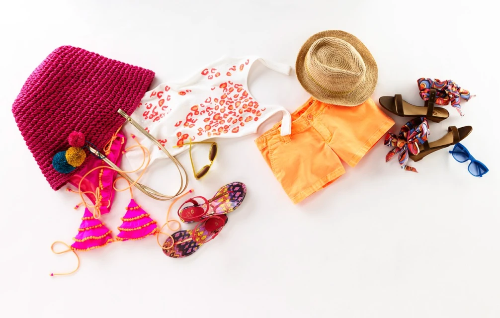 Flat lay of summer fashion and beach accessories including a tote bag, swimsuit, sandals, sunglasses, hat and orange shorts.